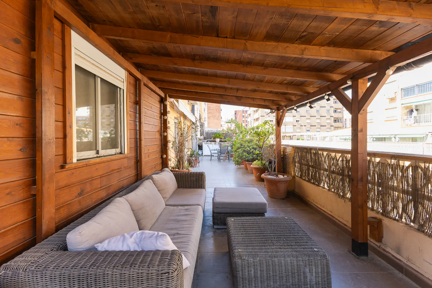 A covered patio with a wicker couch, ottoman, and table. The patio has a wood ceiling and a view of city buildings.