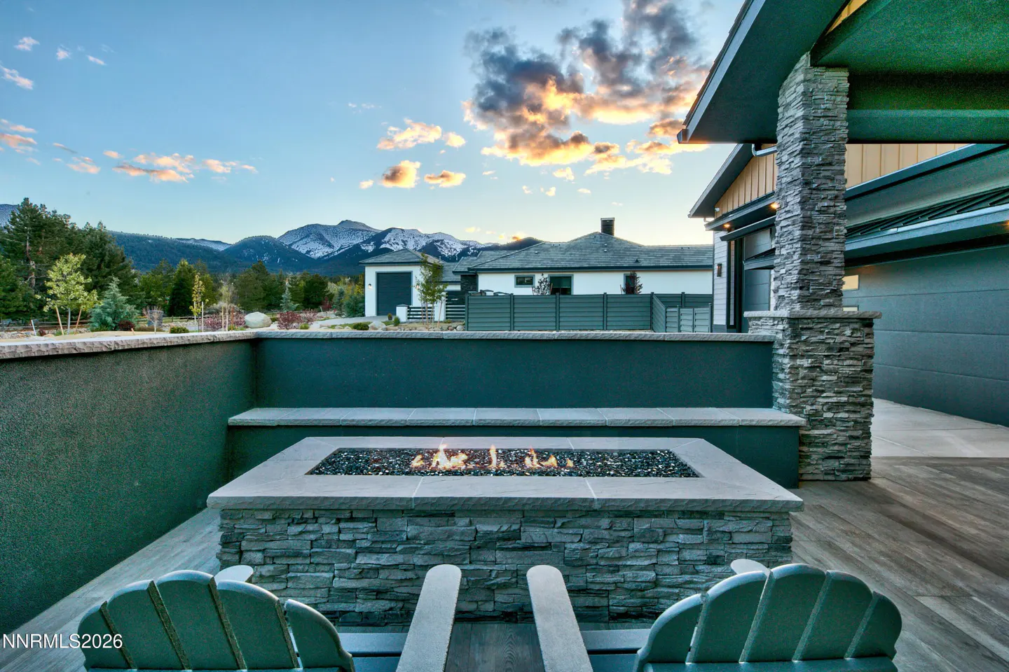 Outdoor patio with stone fire pit and two green Adirondack chairs. Mountains and sunset sky in the background.