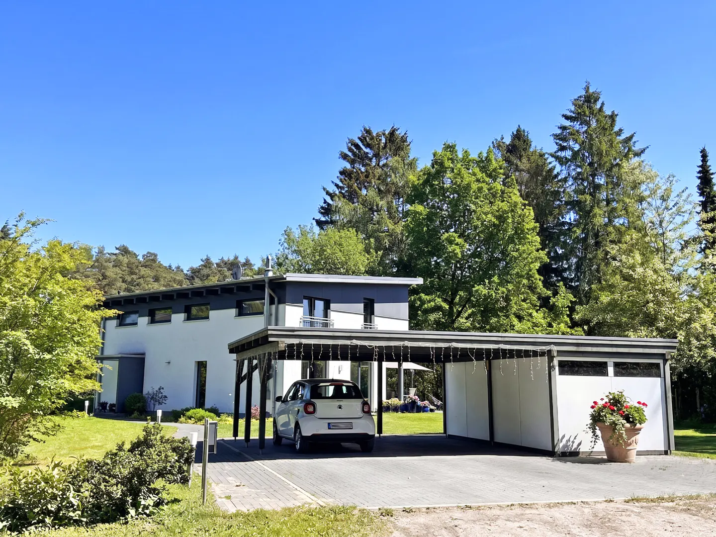 Modern white house with a dark grey roof and a matching carport containing a white car, set in a lush, sunny garden.