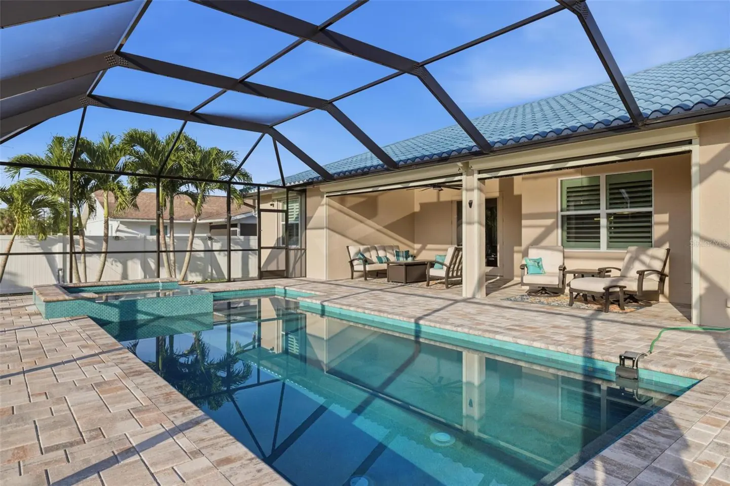 Screened-in pool with blue tile and brick patio. Outdoor seating area with beige furniture and blue accents. Palm trees and blue sky visible.