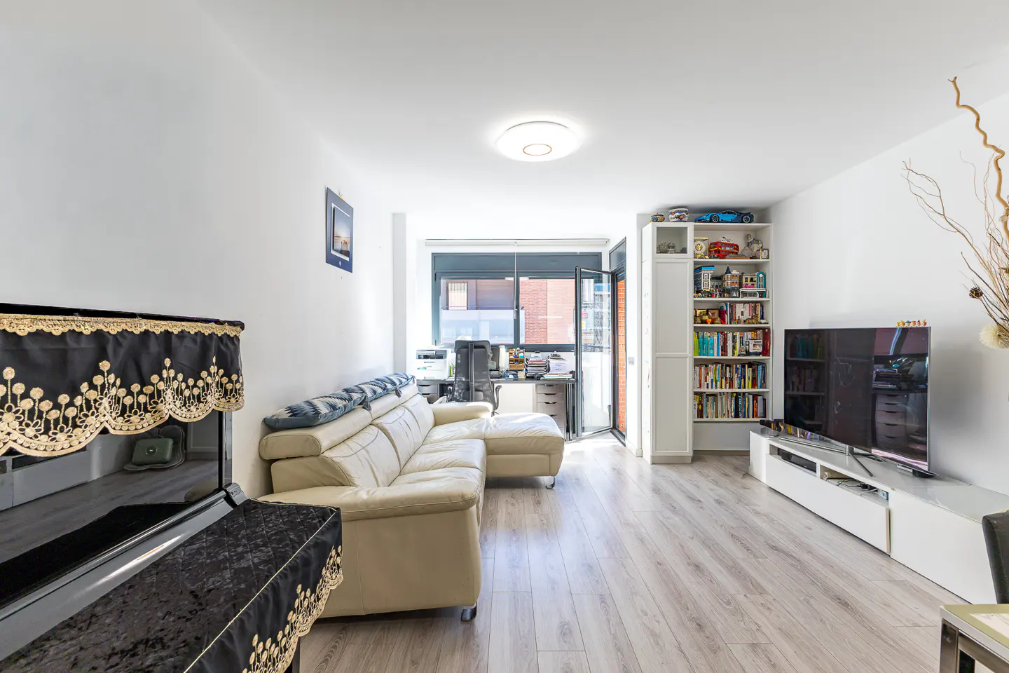 Bright living room with a piano, white sectional sofa, TV, bookcase, and desk near an open balcony door.
