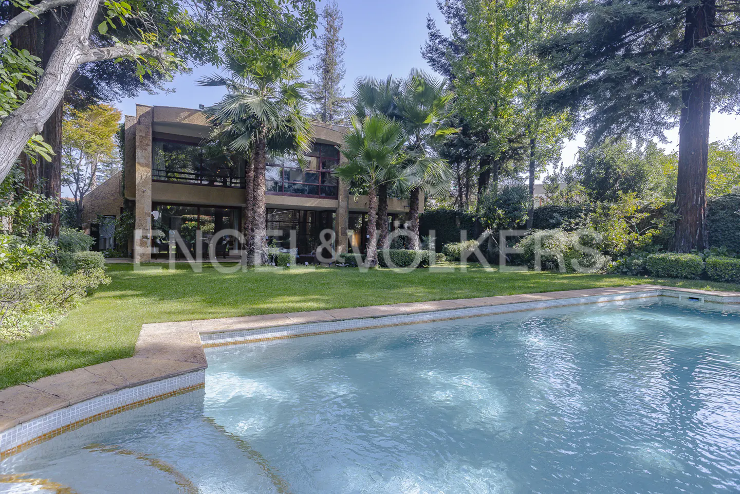 A modern house with a pool and lawn, surrounded by trees. The Engel & Volkers logo is superimposed on the lawn.