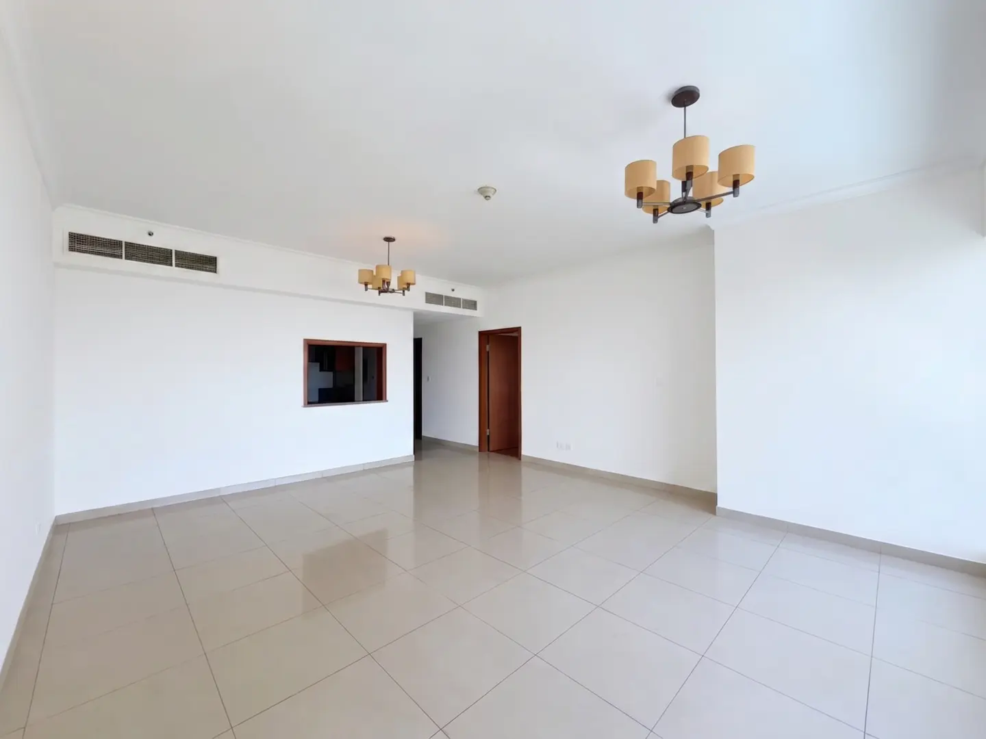 Bright, empty room with white walls, beige tile floor, and two chandeliers. A doorway and small window are visible.
