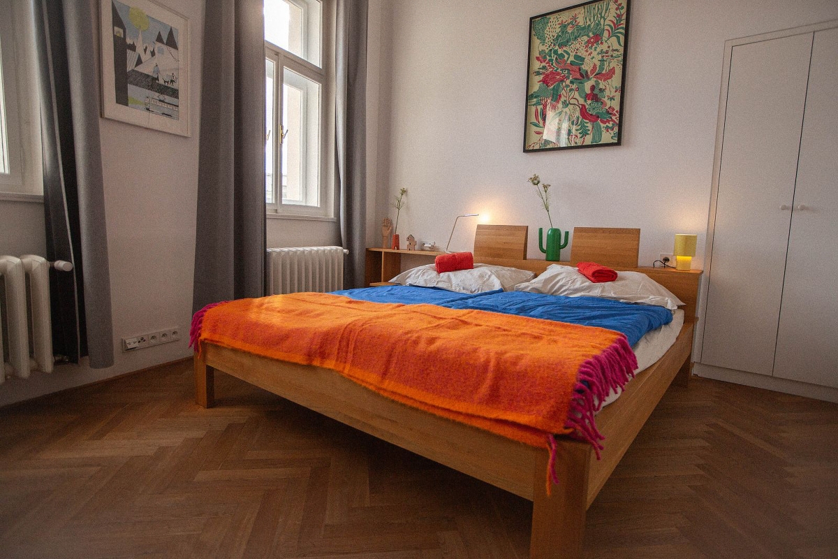 Bright bedroom with a wooden bed, blue sheets, and an orange blanket. Artwork hangs on the white walls. Windows with gray curtains let in natural light.