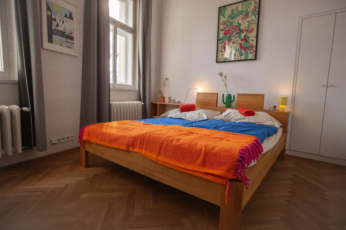 Bright bedroom with a wooden bed, blue sheets, and an orange blanket. Artwork hangs on the white walls. Windows with gray curtains let in natural light.