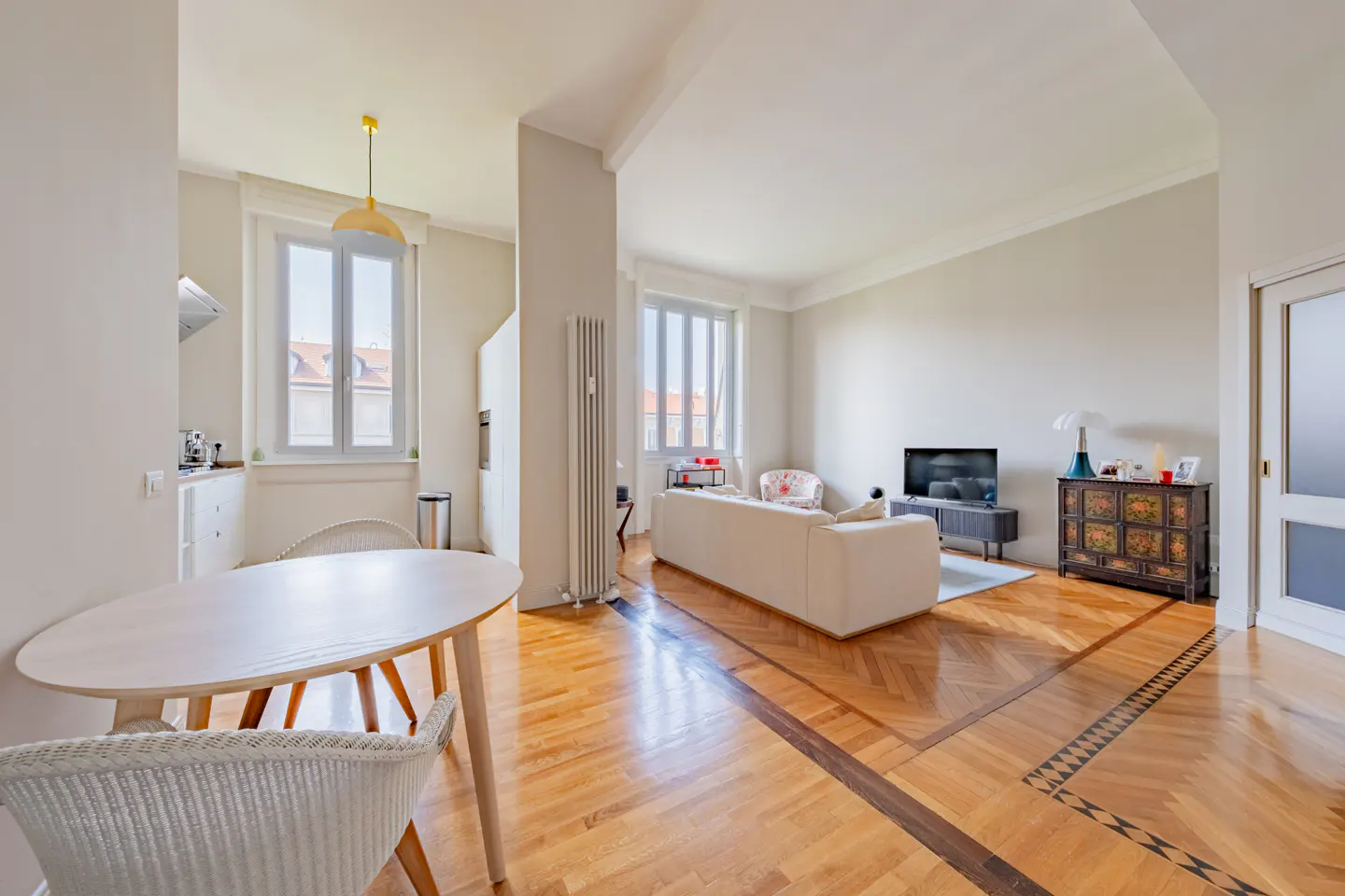 Bright, open-concept apartment with hardwood floors. A white sofa faces a TV. A round table and chairs are in the foreground.