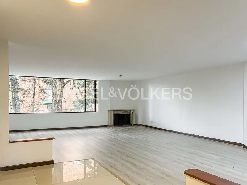 Bright, empty living room with light wood floors, white walls, and a large window showing trees. A fireplace is centered on the far wall.