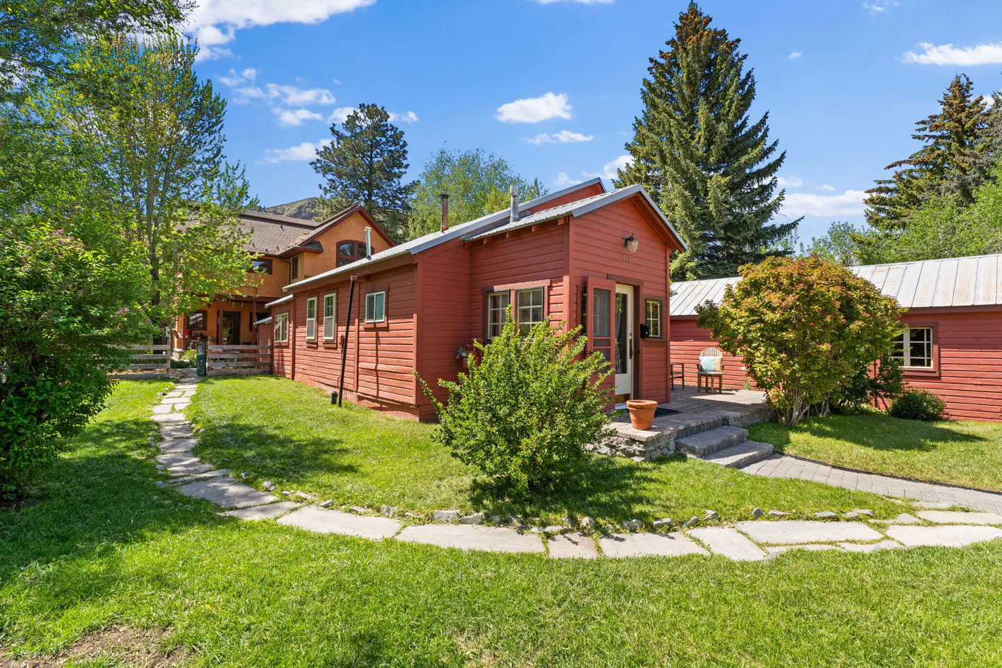 Exterior view of a red wooden house with a stone path leading to the entrance. Green lawn and trees surround the house under a blue sky.