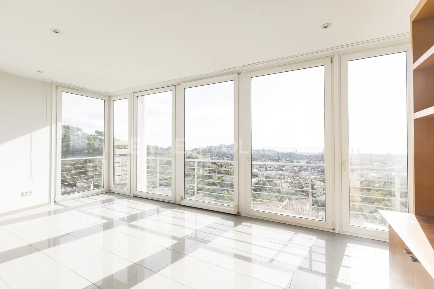 Bright, empty room with white tile floor and large windows. A city view is visible through the windows. A light wood cabinet is on the right.