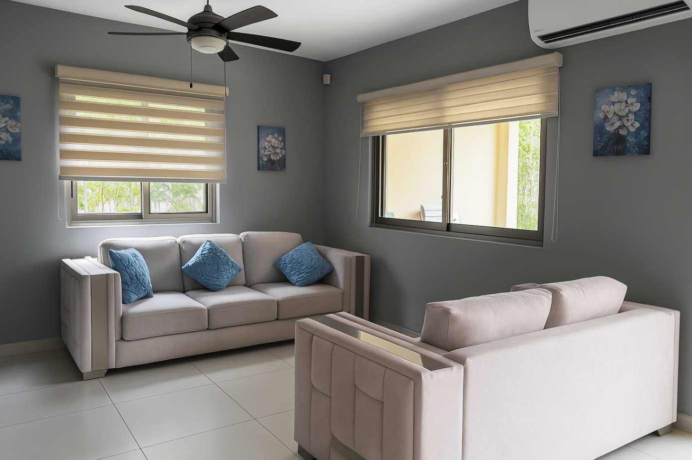 Living room with gray walls, tile floor, and two beige sofas with blue pillows. Windows with blinds, ceiling fan, and wall art complete the space. [AI-generated text]
