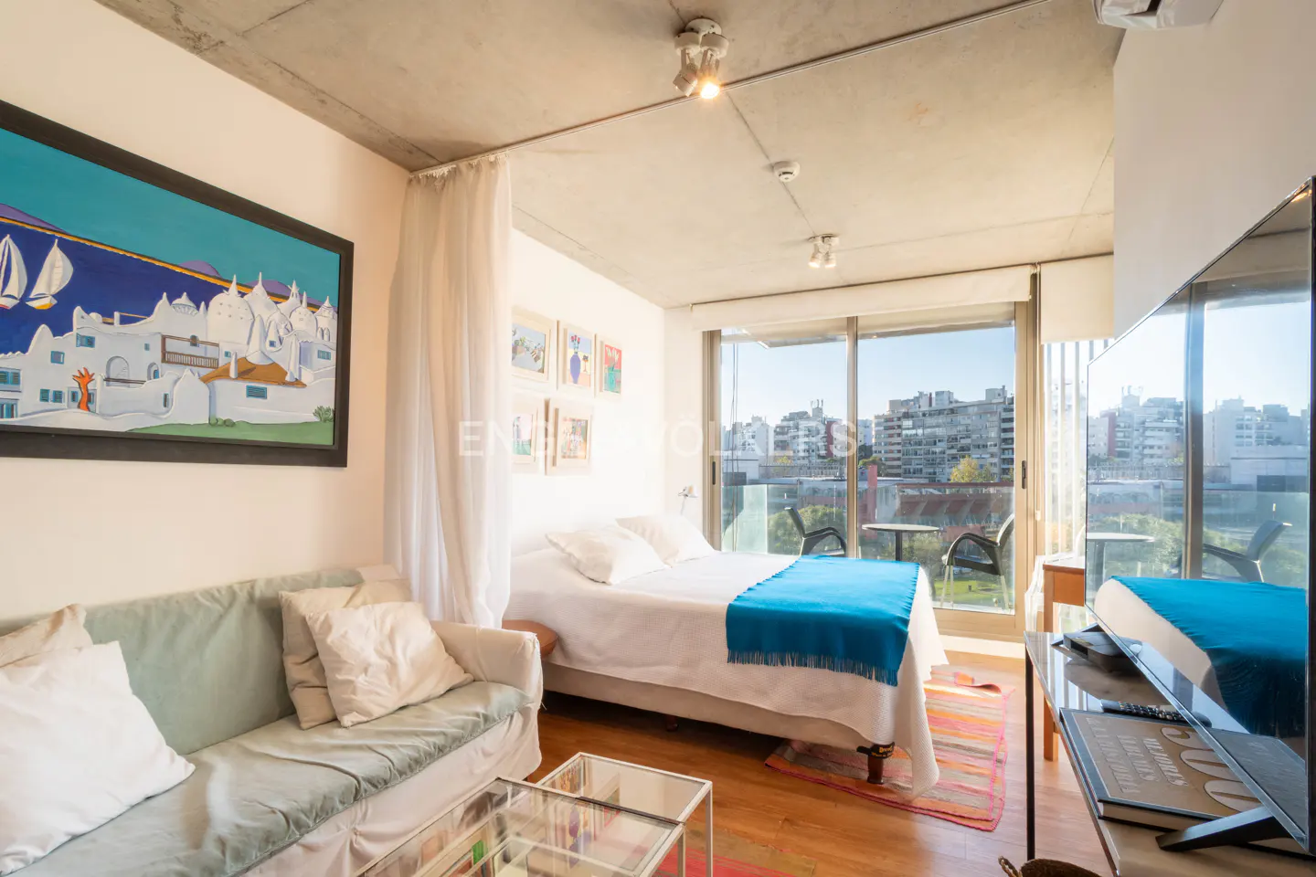 Bright studio apartment with a bed, sofa, and balcony overlooking a city. A large painting hangs on the wall.