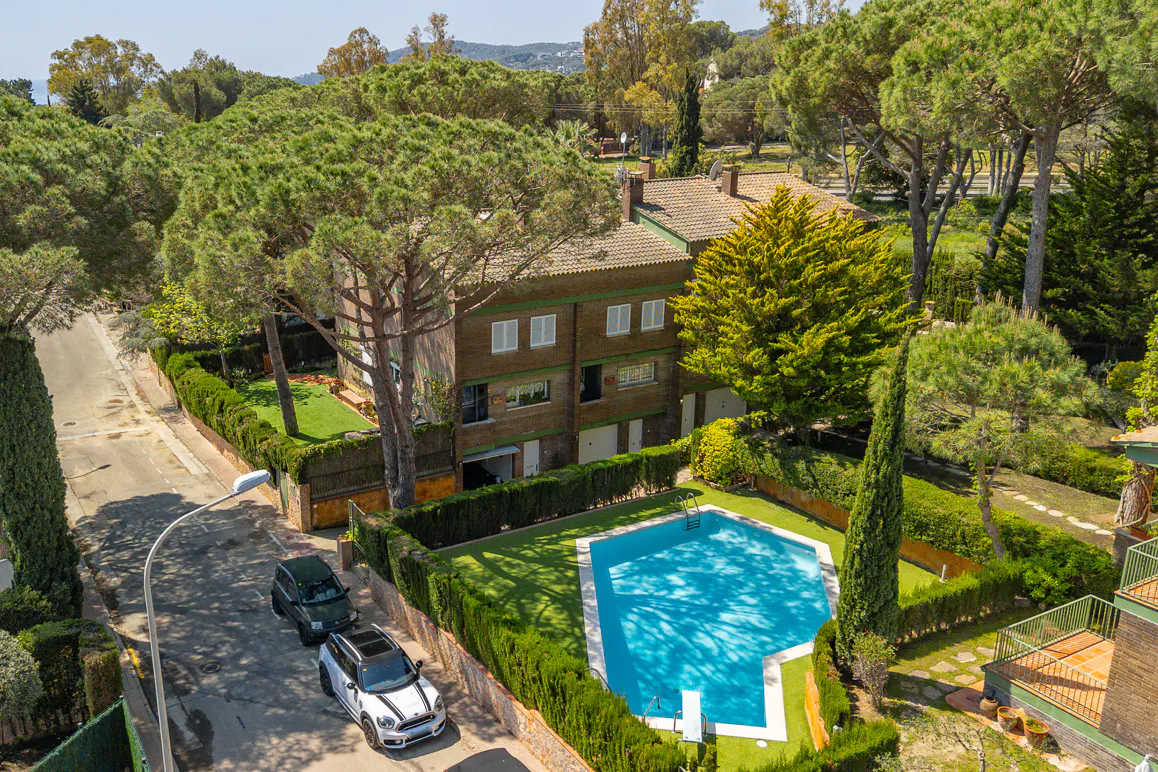 Aerial view of a brick house with a blue pool, green lawn, trimmed hedges, and cars parked on the street.