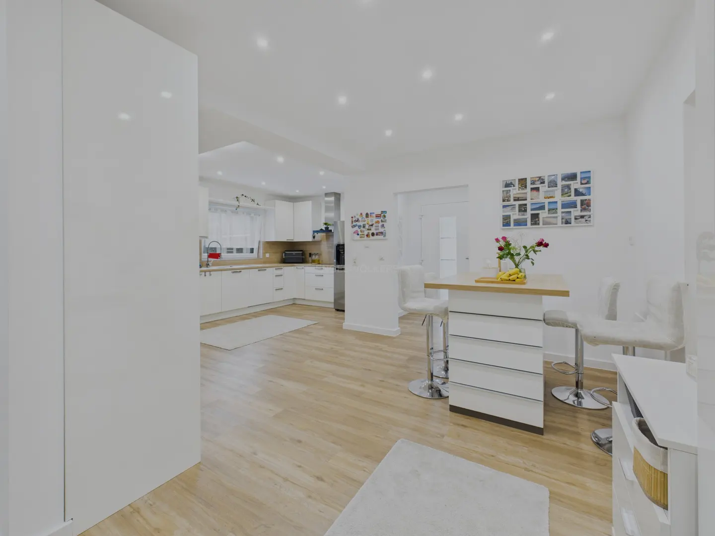 Bright, modern kitchen with white cabinets, wood floors, and a breakfast bar with stools. A collage of photos decorates the wall.