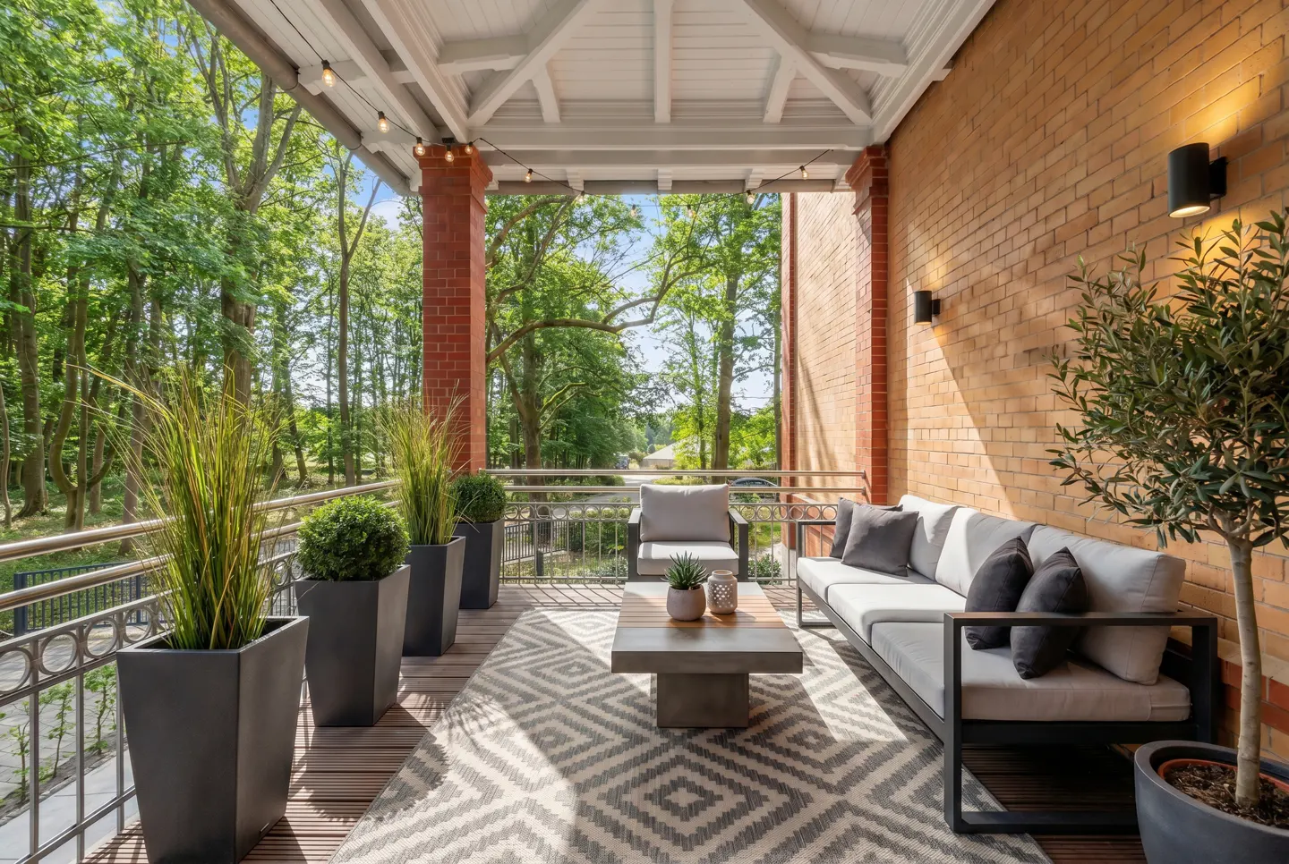 Outdoor patio with gray sofa, patterned rug, and potted plants. Brick wall and trees in the background.