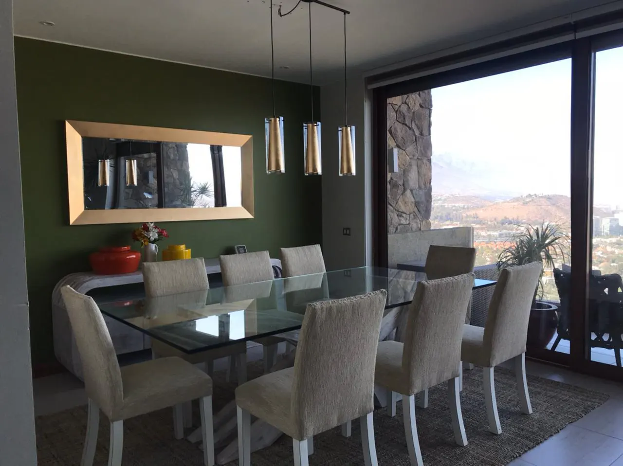 A dining room with a glass table, beige chairs, and a large window with a view. The walls are olive green, and a gold-framed mirror hangs on the wall.