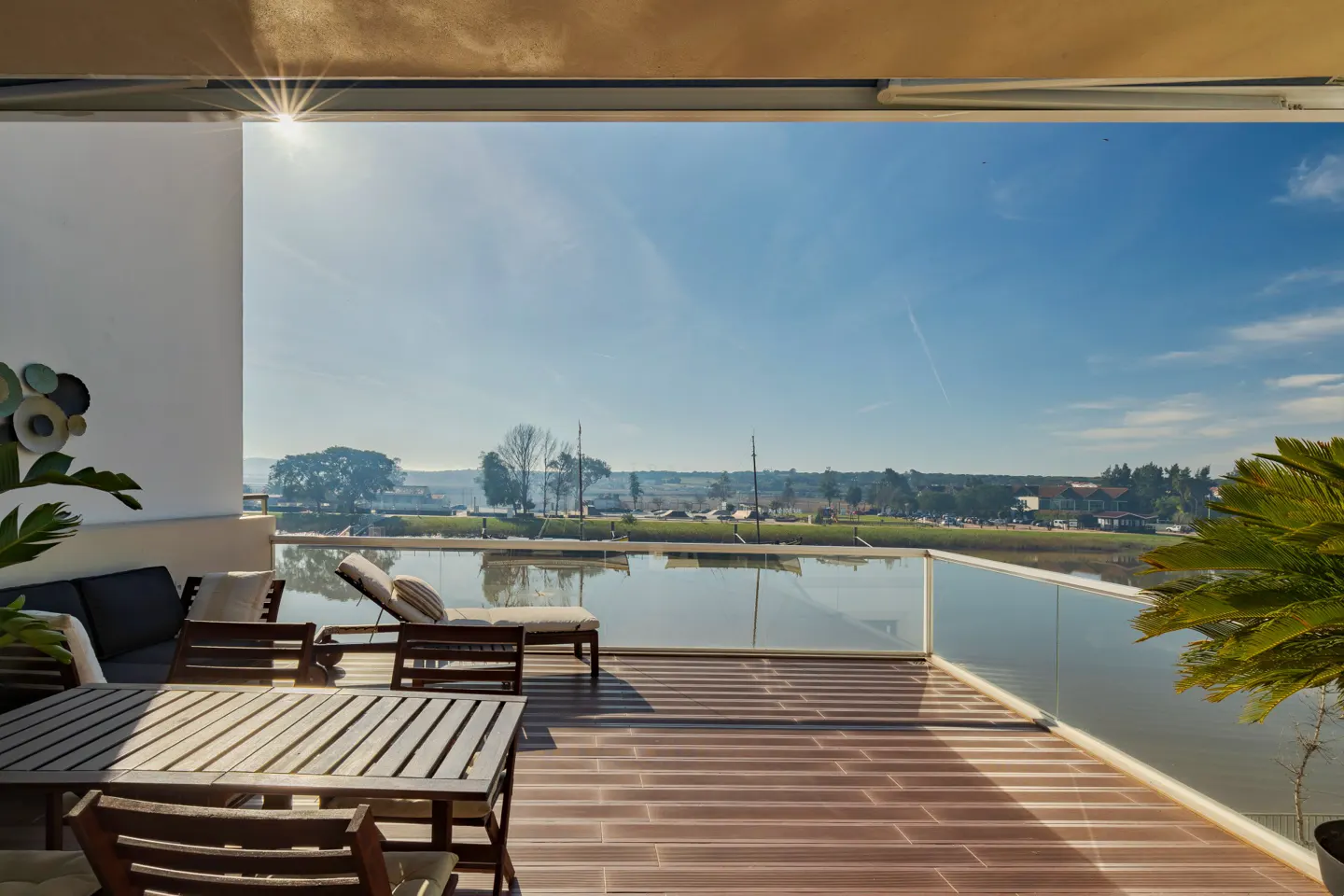 Sunny balcony with wooden deck, outdoor dining set, and lounge chair overlooking a calm river and distant landscape.