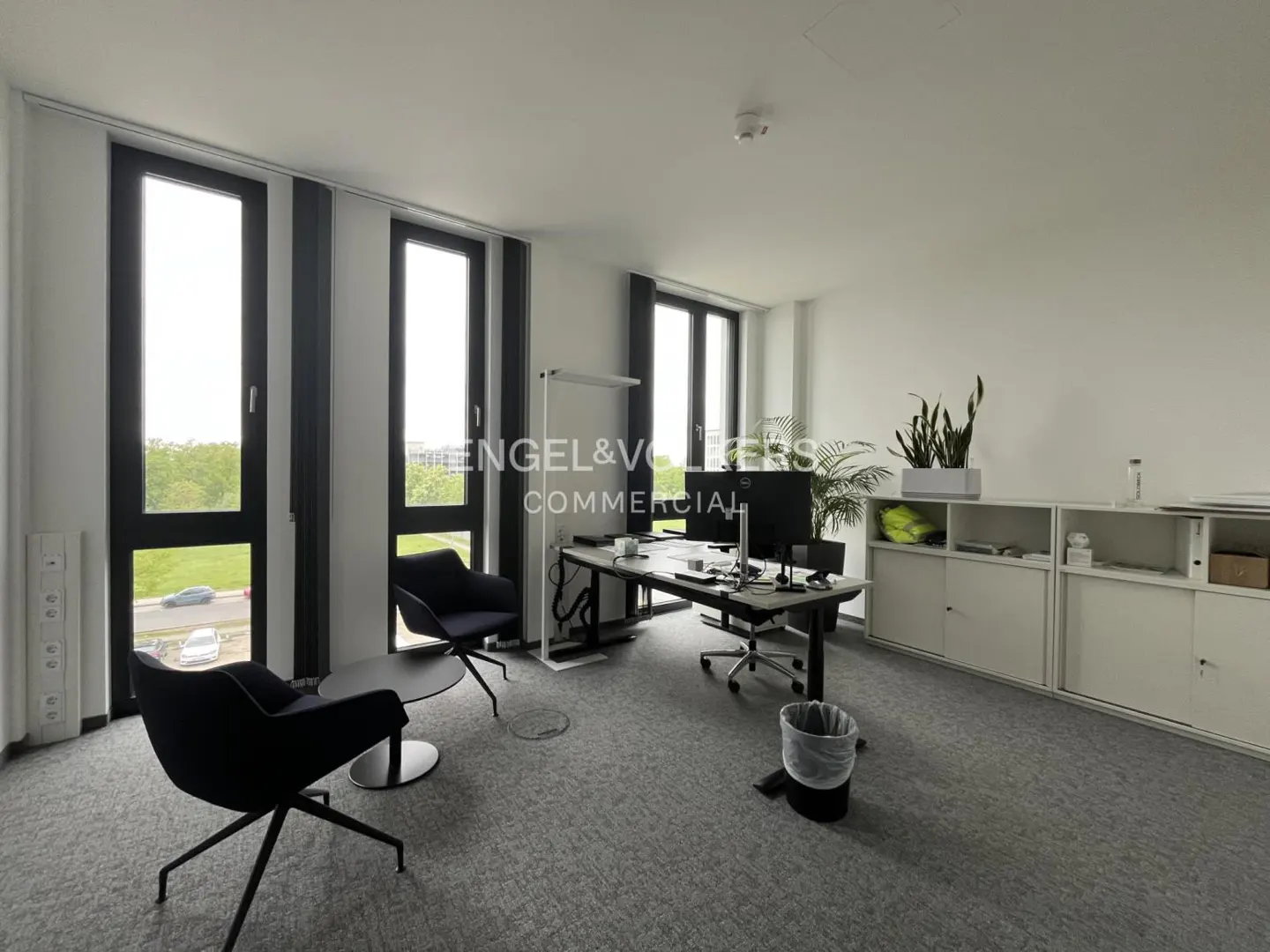 Bright office space with gray carpet, black chairs, a desk with computers, and white cabinets. Large windows offer a view of greenery.