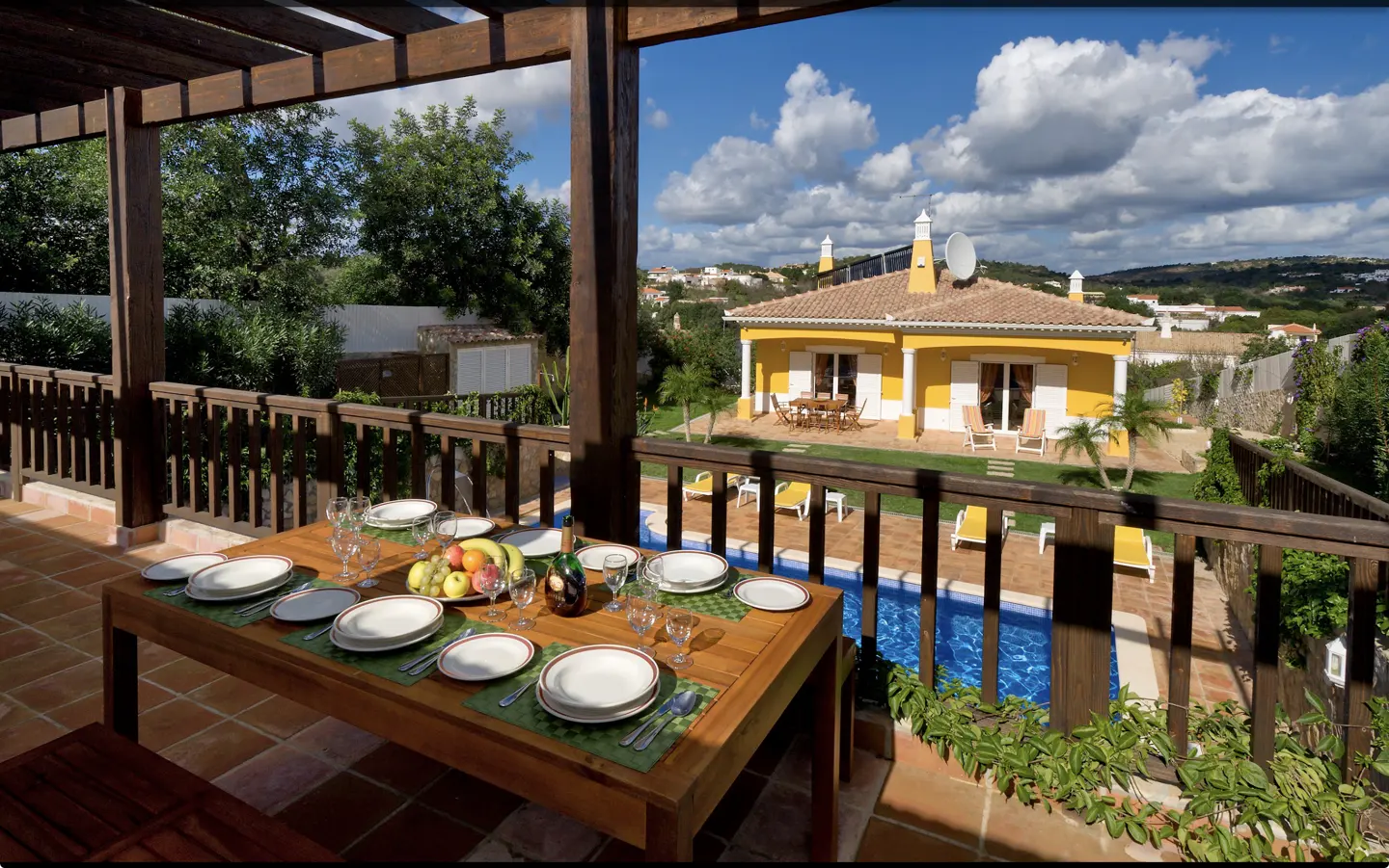 A wooden dining table on a covered terrace overlooking a yellow villa with a swimming pool and garden under a blue sky.