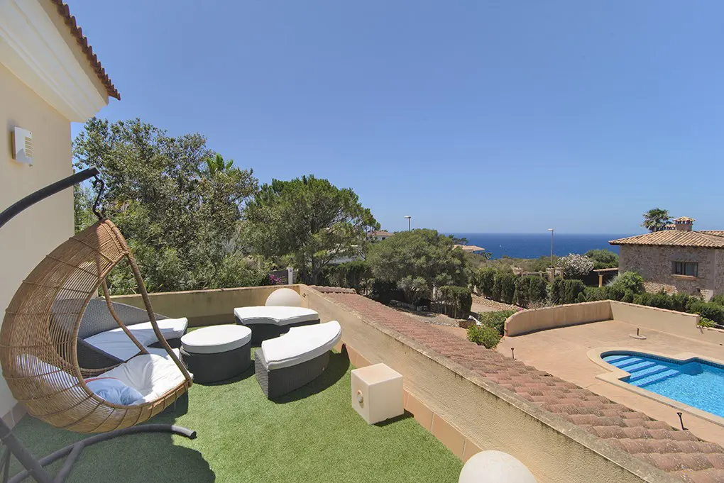 Rooftop terrace with a hanging wicker chair, lounge seating, and artificial grass, overlooking a pool and the blue sea.