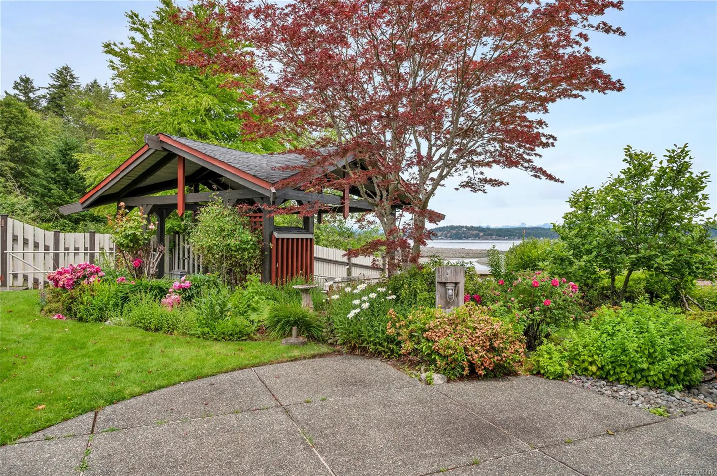 Landscaped yard with a dark wood gazebo, red maple tree, flowers, and a view of the ocean in the background.