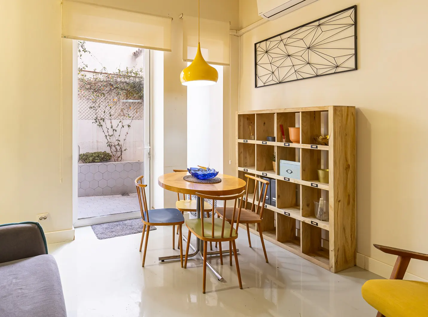 Bright dining area with a round wooden table, colorful chairs, and a yellow pendant light. A wooden bookshelf and geometric wall art add character. Patio view through open doors.