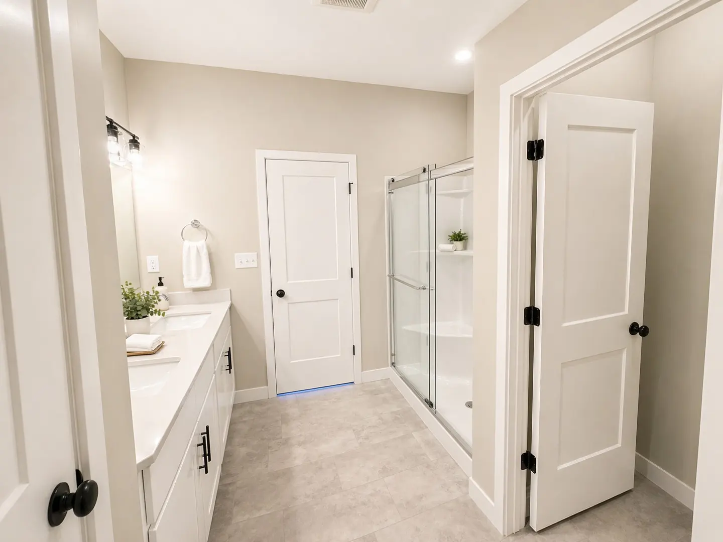 Bright bathroom with gray tile floor, white cabinets, double sink, glass shower, and white doors with black hardware.