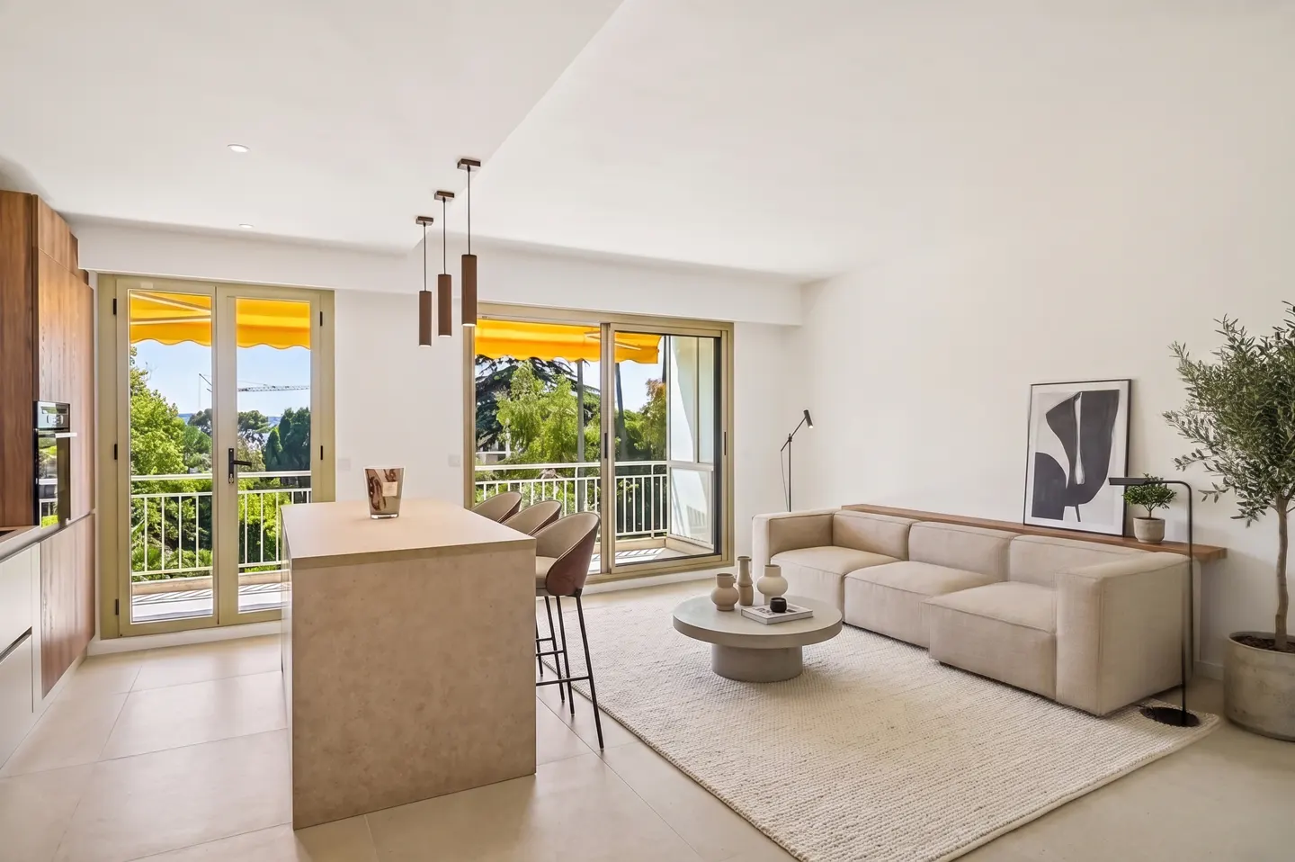 Bright, modern living room with a beige sofa, round coffee table, and kitchen island. Balcony doors with yellow awnings overlook lush greenery.
