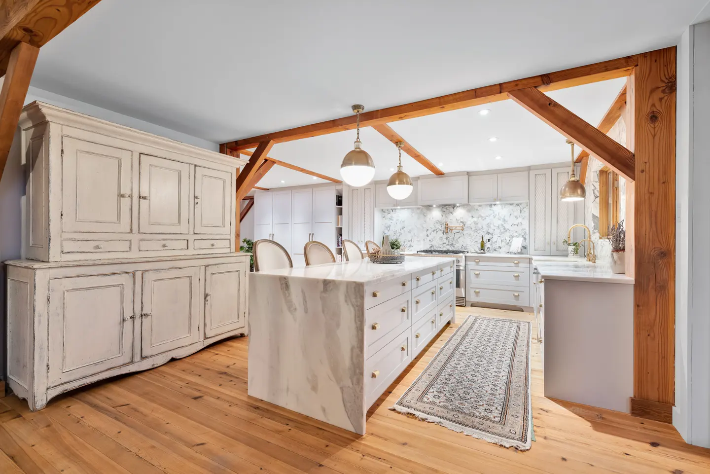 Bright kitchen with wood floors, beams, and a marble island. A white hutch sits to the left.