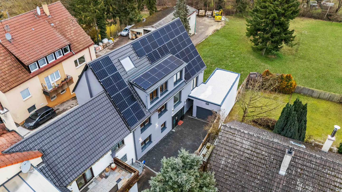 Aerial view of a modern gray house with solar panels on the roof, surrounded by green lawns and neighboring houses.