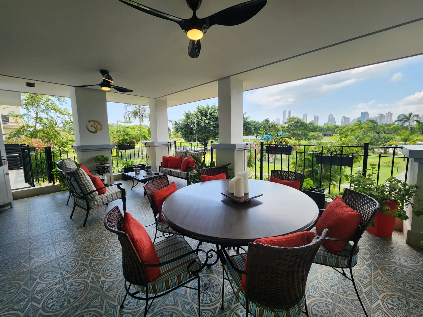 Covered patio with round table, wicker chairs with orange pillows, ceiling fans, and a view of a park and city skyline.