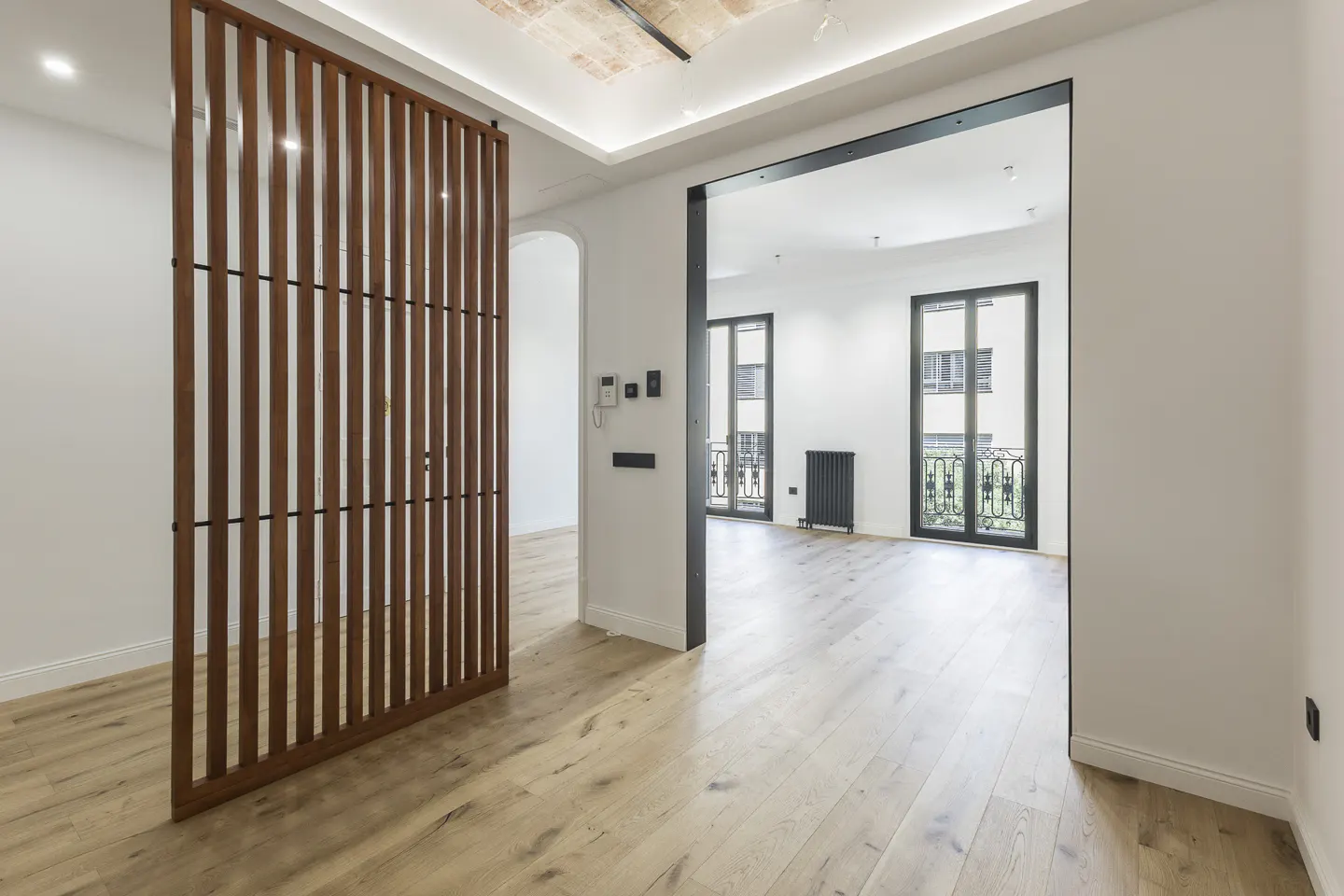 Interior view of a bright, modern apartment with light wood floors, a brown slat wall, and a black-framed doorway to a room with balcony doors.