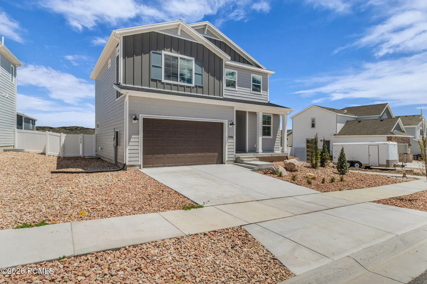 Two-story house with gray siding, brown garage door, and white trim under a blue sky. A concrete driveway leads to the garage. The yard is covered in brown gravel.