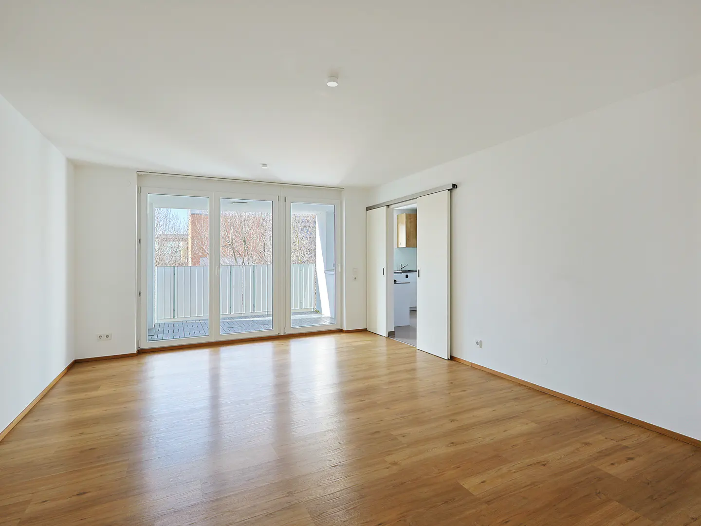 Bright, empty room with wood floors, white walls, and sliding glass doors to a balcony. A sliding door reveals a glimpse of a kitchen.