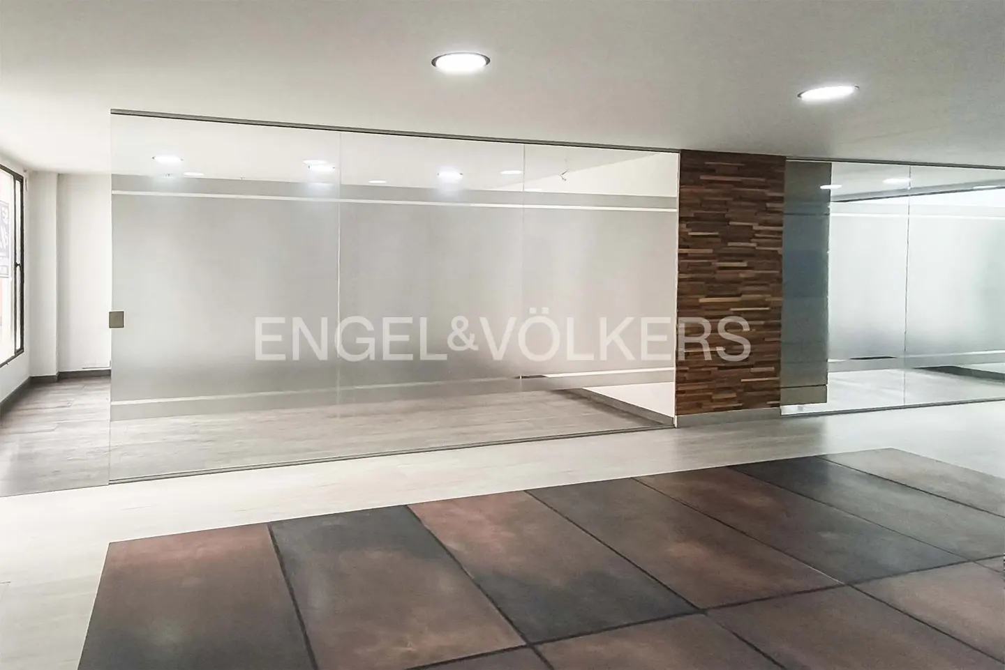 An office space with glass walls, wood paneling, and a brown tiled floor. The Engel & Völkers logo is on the glass.