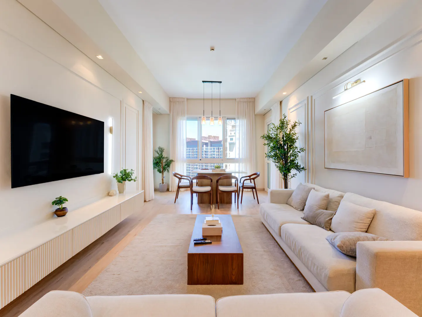 Bright, modern living room with a large TV, white sofas, a wooden coffee table, and a dining area with city views.