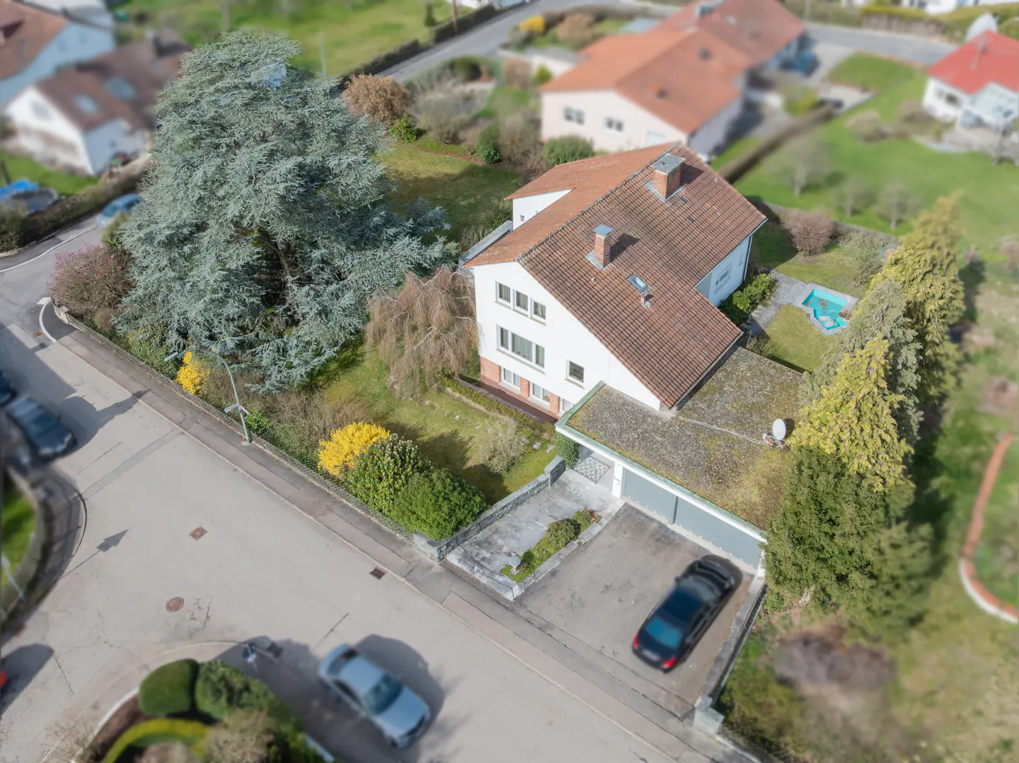 Aerial view of a white house with a brown tiled roof, large tree, garage, and driveway with a car in a suburban setting.