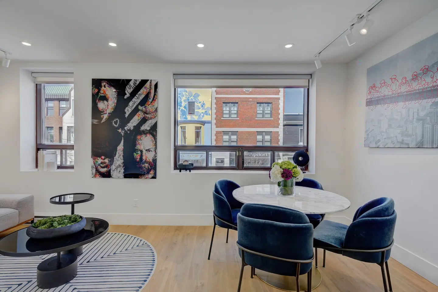 Bright living room with wood floors, white walls, and modern art. A round marble table with blue velvet chairs sits near a window.