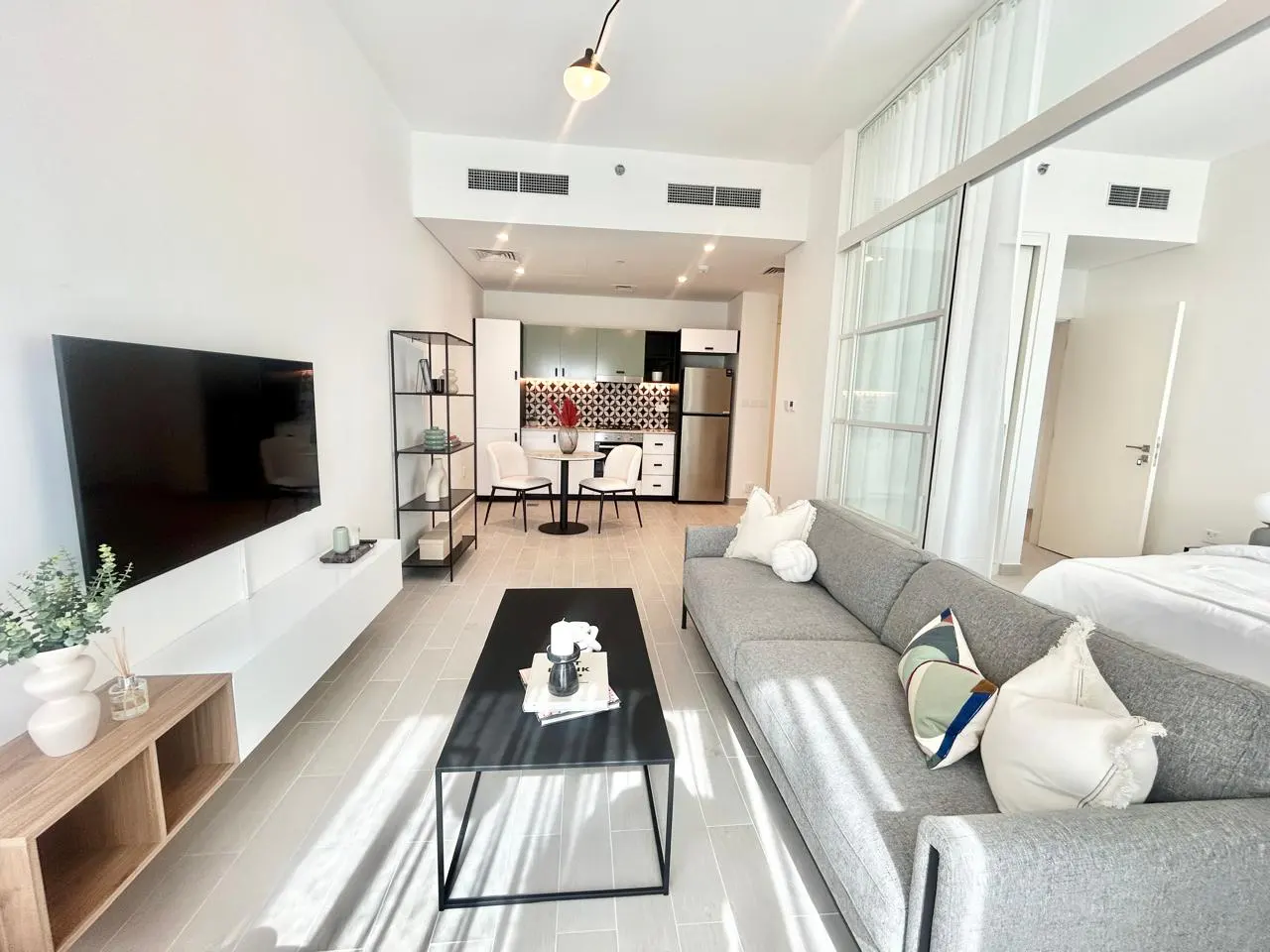 Bright, modern apartment interior with a gray sofa, black coffee table, TV, and open kitchen with white cabinets and stainless steel appliances.