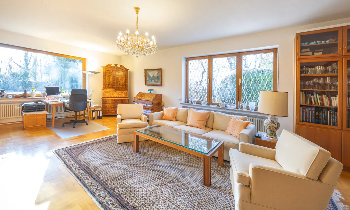 Bright living room with a beige sofa, two armchairs, a glass coffee table, and a large window overlooking trees. A desk and bookcase are also visible.