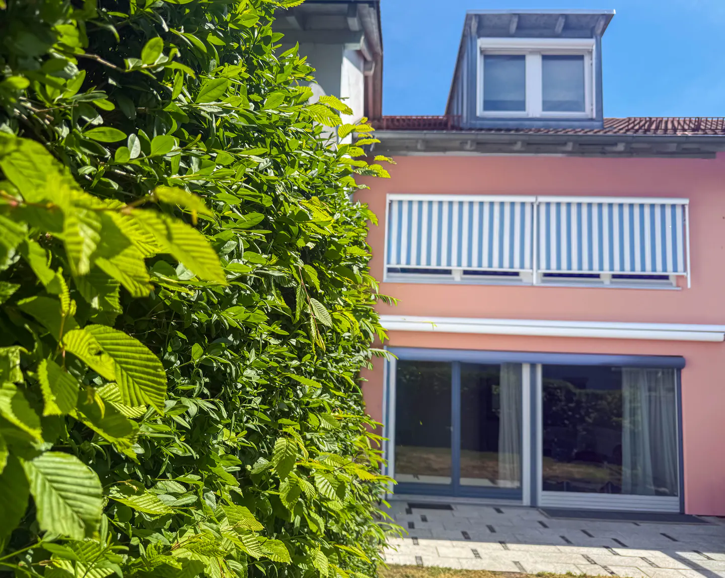 A pink two-story house with blue and white striped awnings, seen behind a green hedge.
