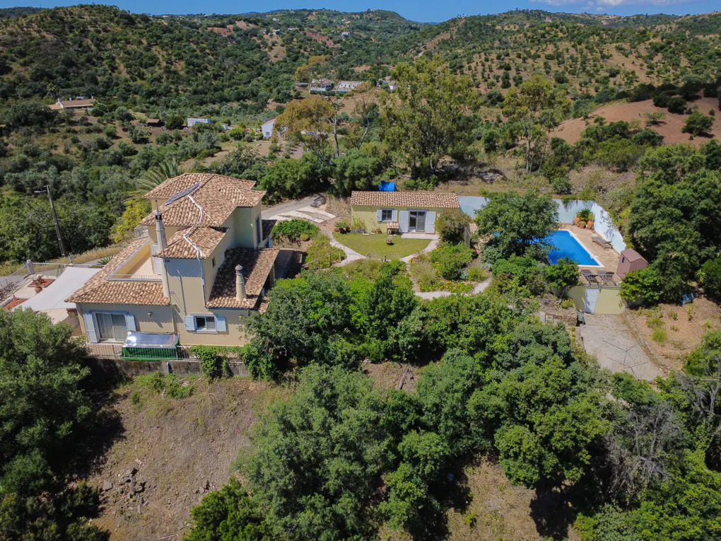 Aerial view of a tan house with a red tile roof, a pool, and green trees in a hilly area.