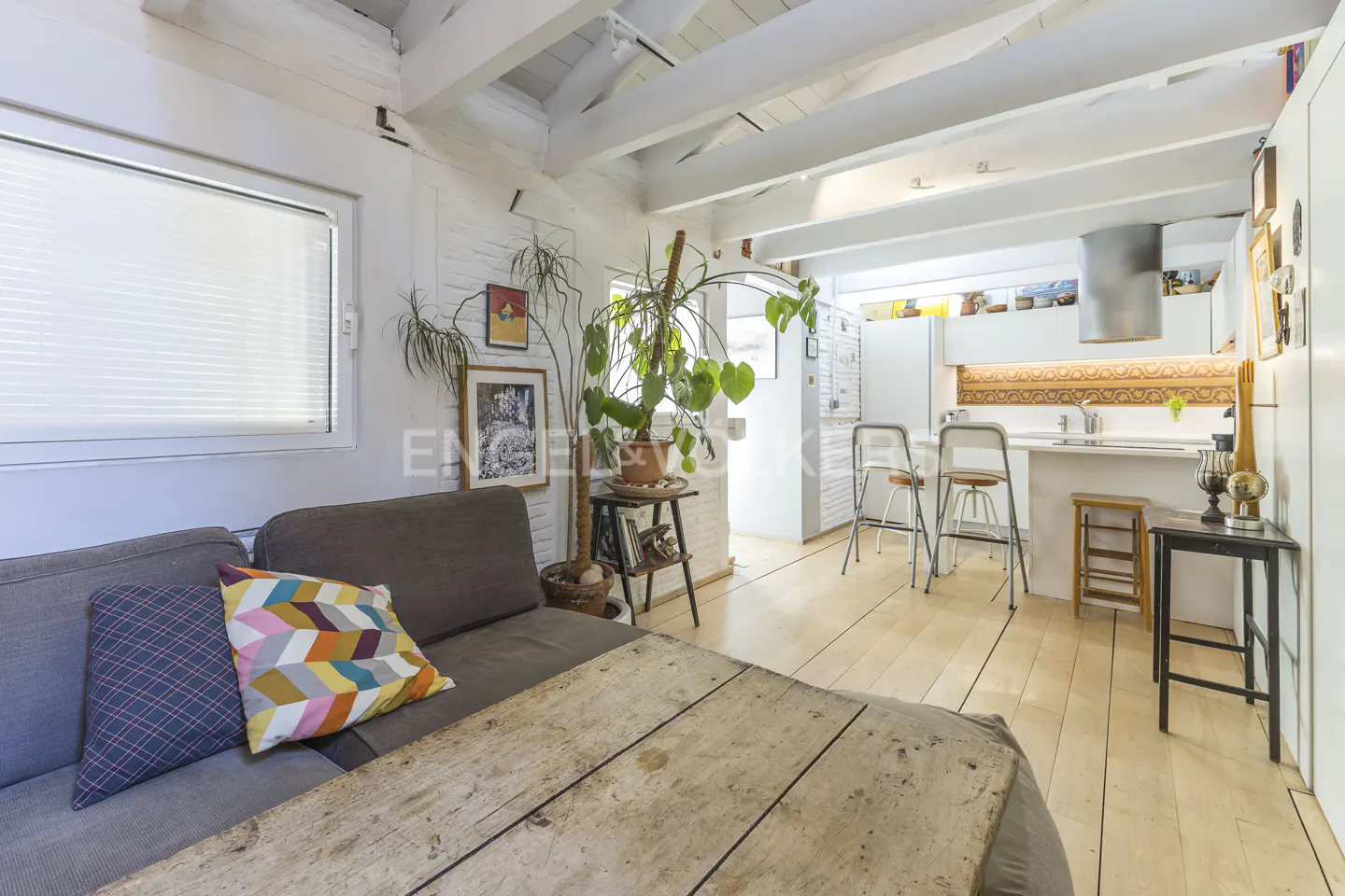Bright, open-plan living space with white walls, wooden beams, and light wood floors. A gray sofa with colorful pillows sits near a rustic wooden table. A kitchen with bar stools is visible in the background.
