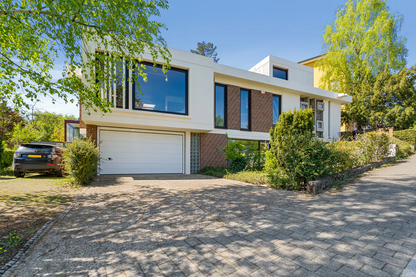 Modern two-story house with white siding, brick accents, and black-framed windows. A white garage door faces a paved driveway. Green trees surround the property.