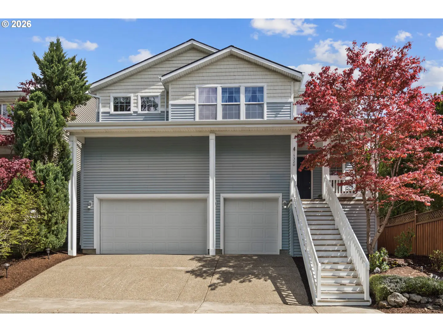 Two-story townhouse with gray siding, white trim, and two garage doors. A red-leaved tree stands to the right of the entrance stairs.