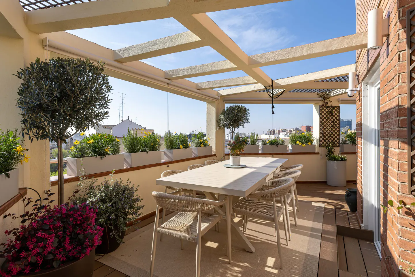 Outdoor dining area on a rooftop terrace with a long white table, rope chairs, potted plants, and a pergola. Brick wall and city view in the background.