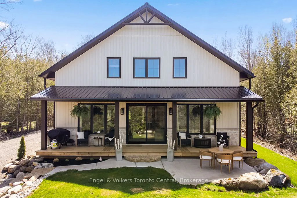 A two-story white farmhouse with a black roof and porch. A patio with a table and chairs is in front.
