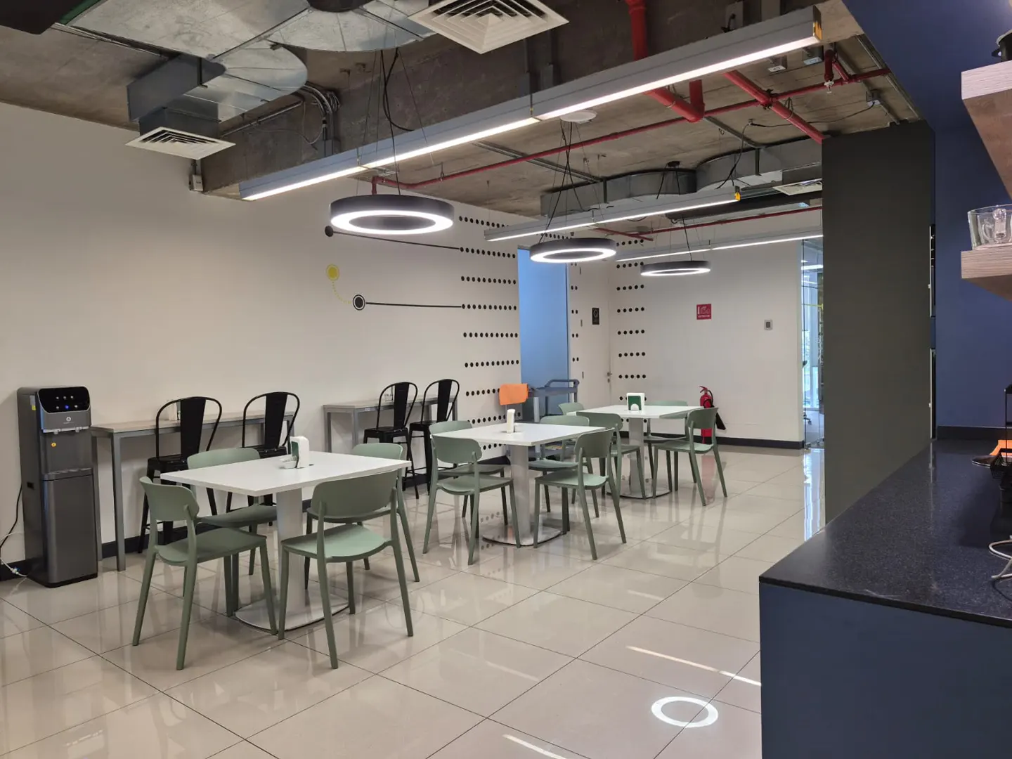 A modern office break room with white tables, green chairs, and round pendant lights.