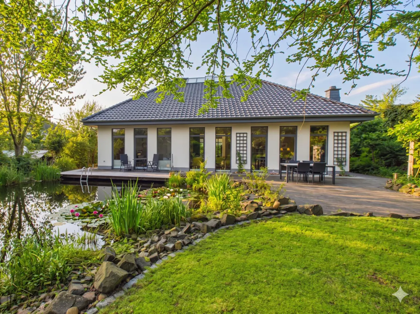 A modern white house with a gray roof overlooks a pond with lily pads and a green lawn.