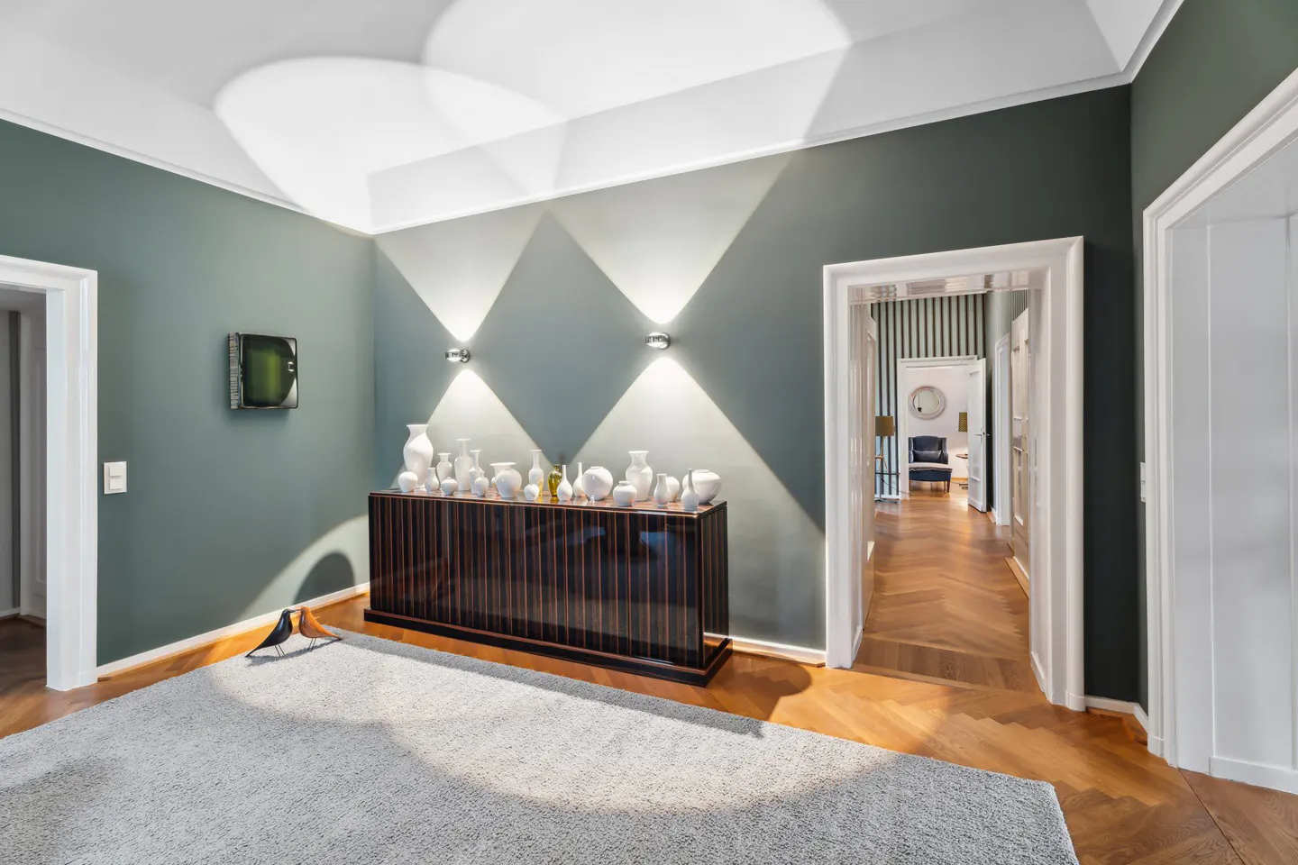 A room with green walls, a dark wood cabinet topped with white vases, and a gray rug. Two wall sconces cast light in a diamond pattern.