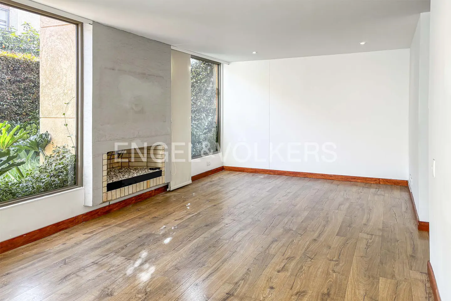 Bright, empty living room with wood floors, white walls, and a brick fireplace. Large windows show greenery outside.